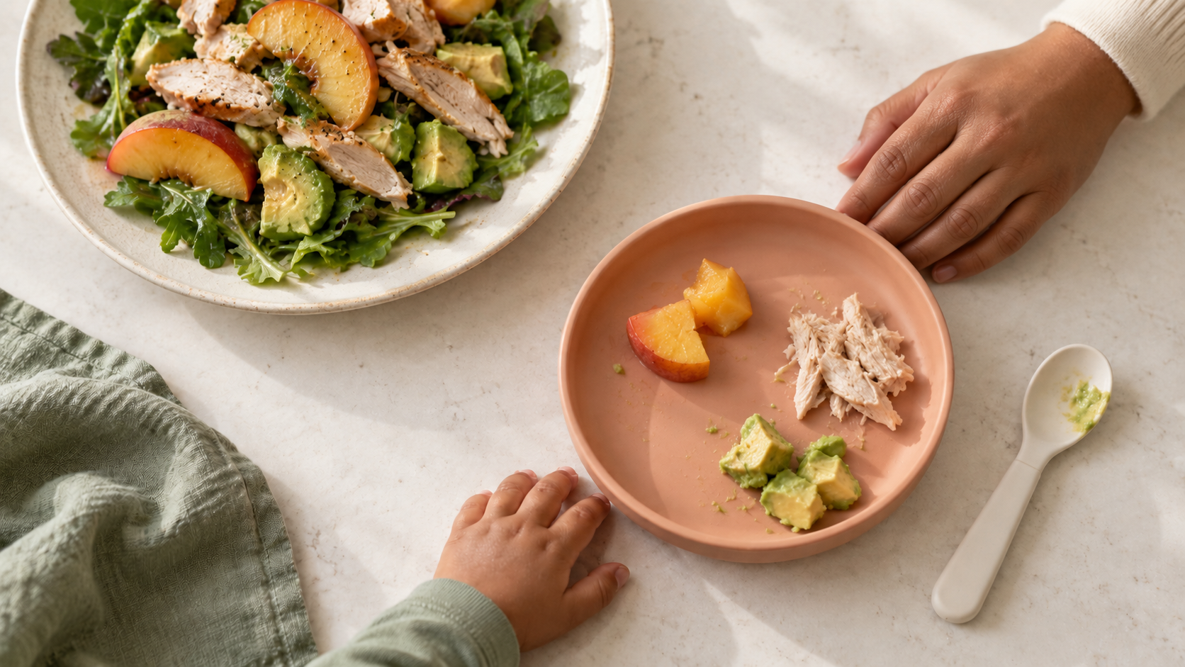 A caregiver and child pause beside a small plate of peaches, chicken, and avocado